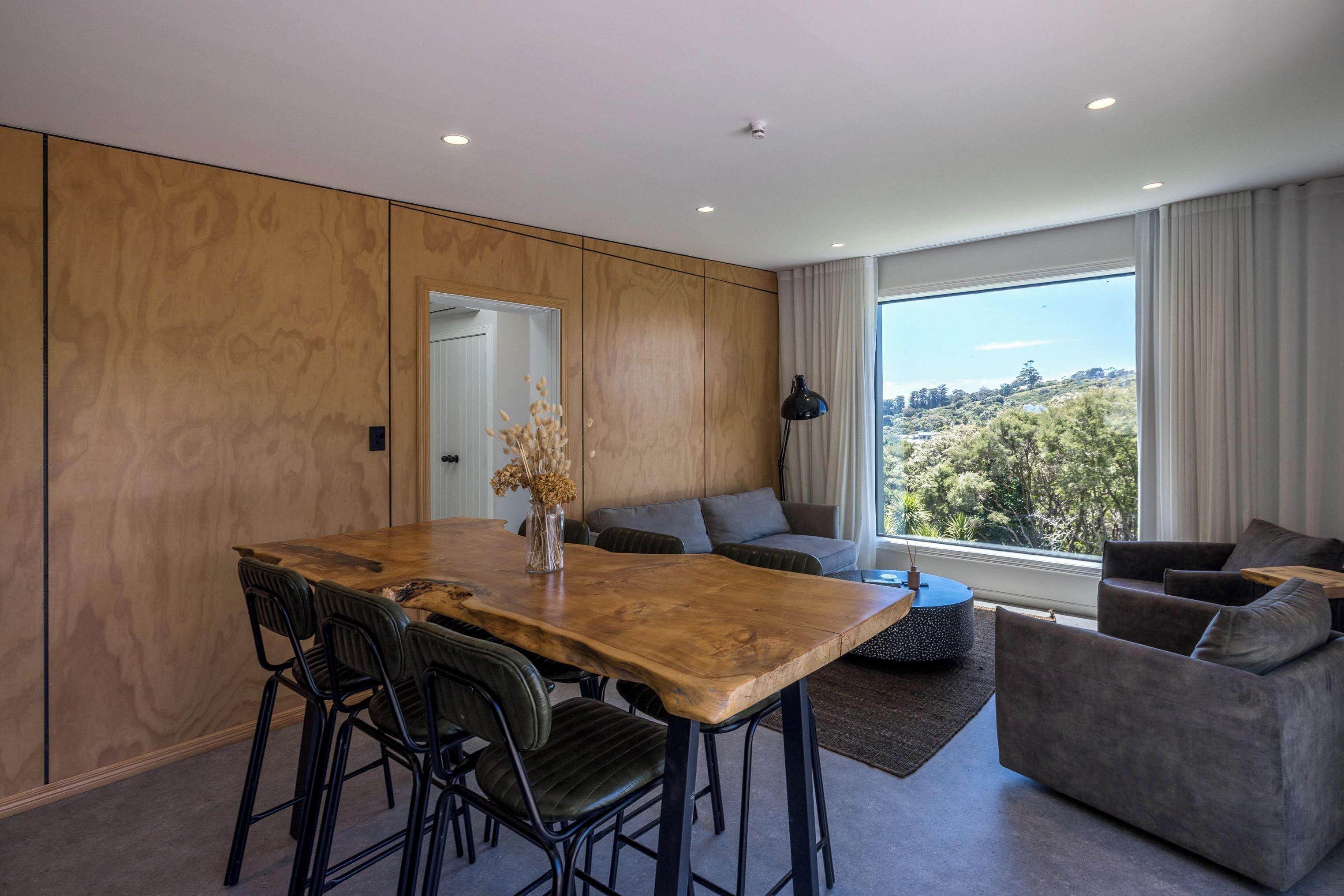 A modern living room with a wooden dining table, grey sofas, and a large window offering a view of trees.