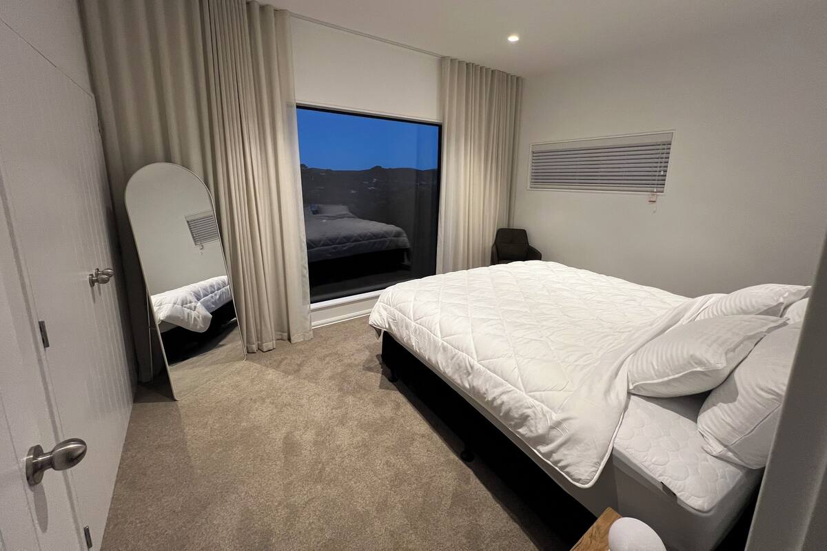 A modern hotel room with a large bed, a window with curtains, a bedside table, and a mirror.