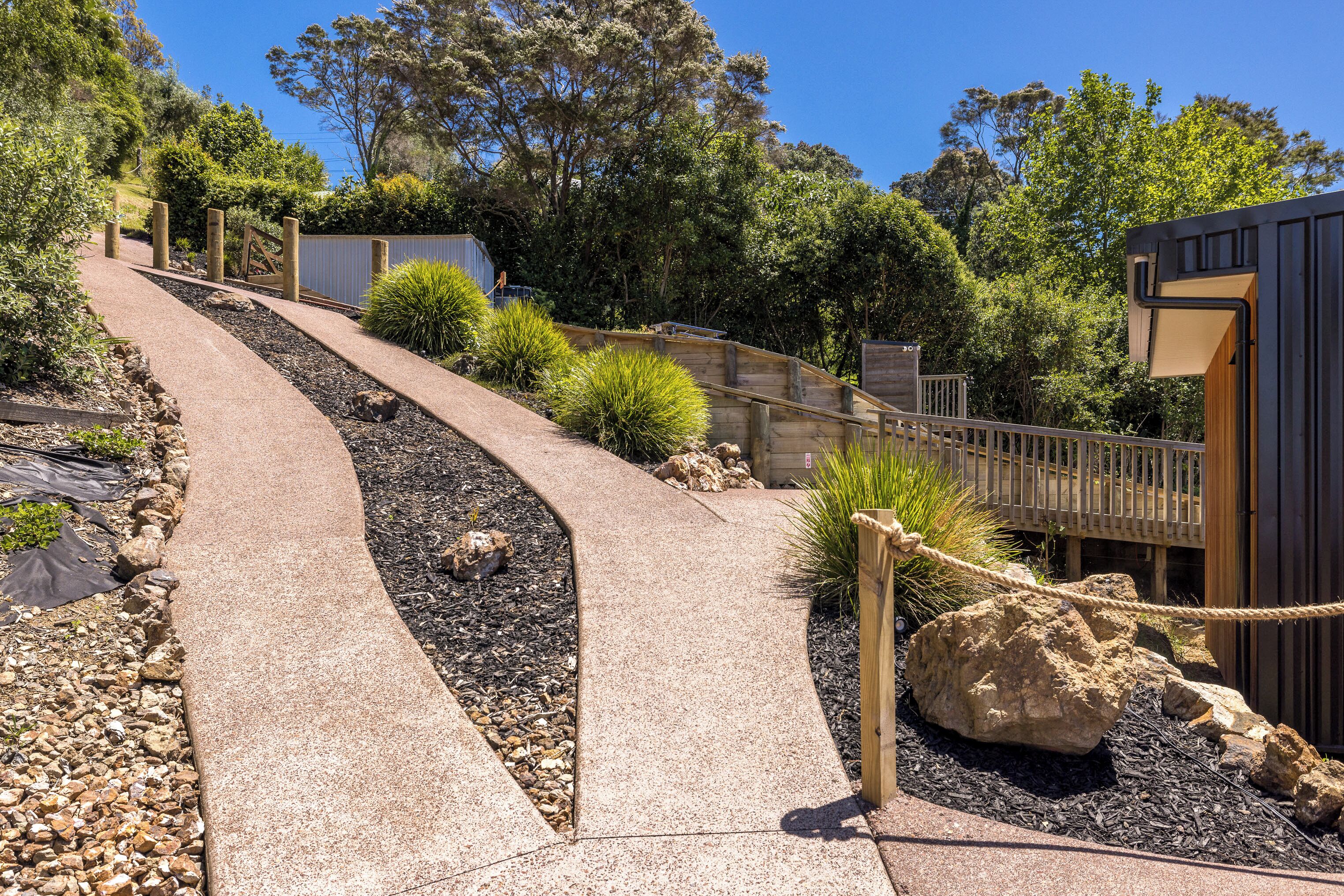 A concrete pathway leading to a wooden deck with a rope barrier, surrounded by rocks and plants.