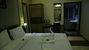 Habitación de hotel con cama bien hecha, armario, escritorio con hervidor de agua y baño con lavamanos y espejo.