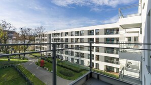 Un edificio moderno de varios pisos con un balcón de vidrio, un patio ajardinado y un cielo despejado.