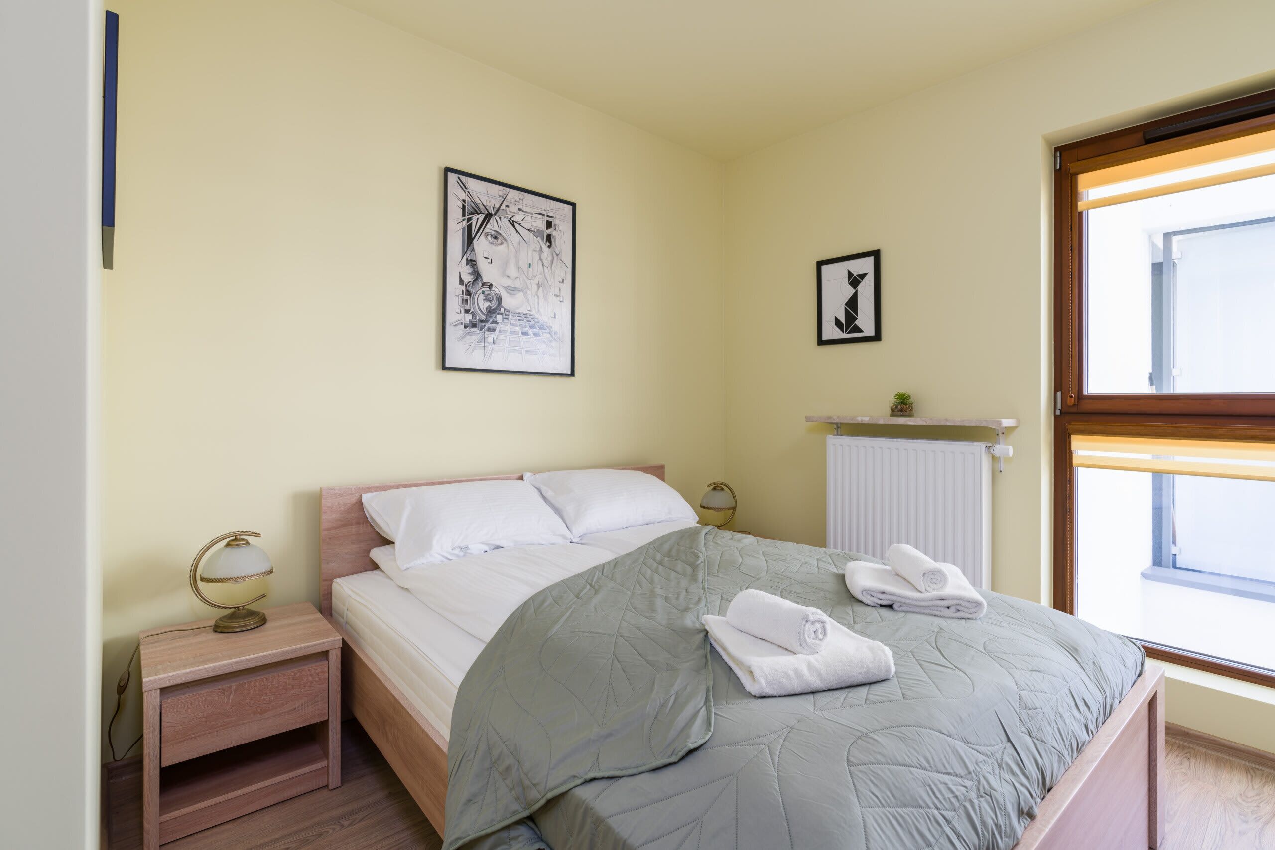 Un lit bien fait, avec une couverture grise et des oreillers blancs, une lampe de chevet et un tableau encadré au mur.