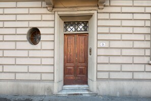 Une porte en bois à deux battants, surmontée d’une grille métallique, est encastrée dans un mur en briques de couleur beige, avec une petite fenêtre circulaire sur le côté gauche.