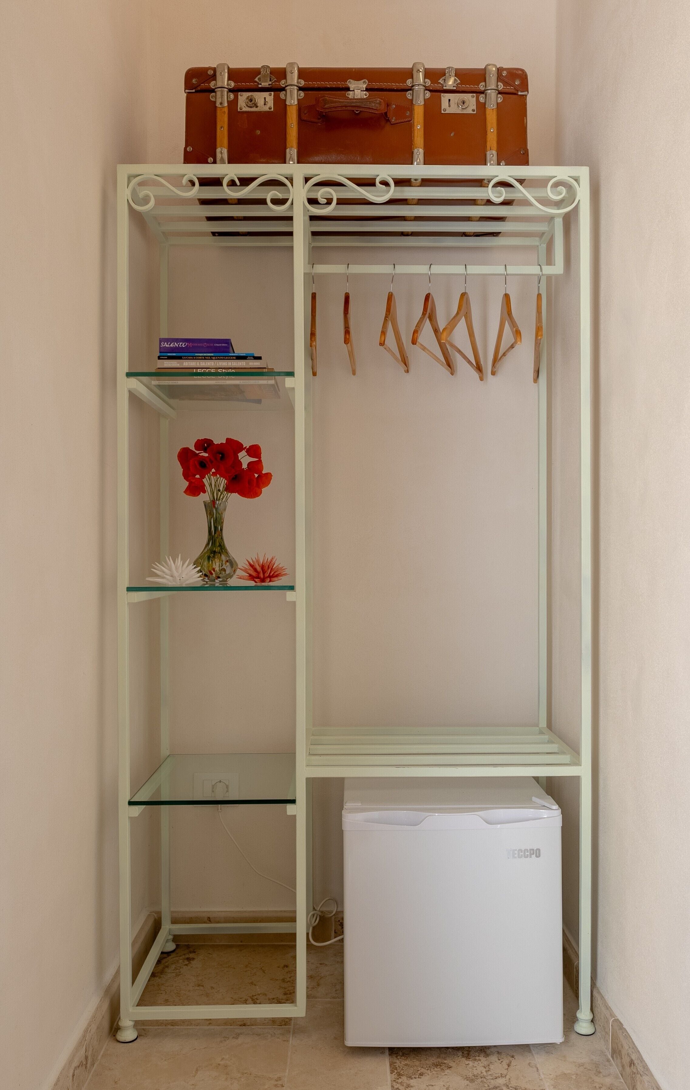 Ein weißes Metallregal mit einem Koffer obenauf, einem kleinen Kühlschrank und einer Vase mit roten Blumen.