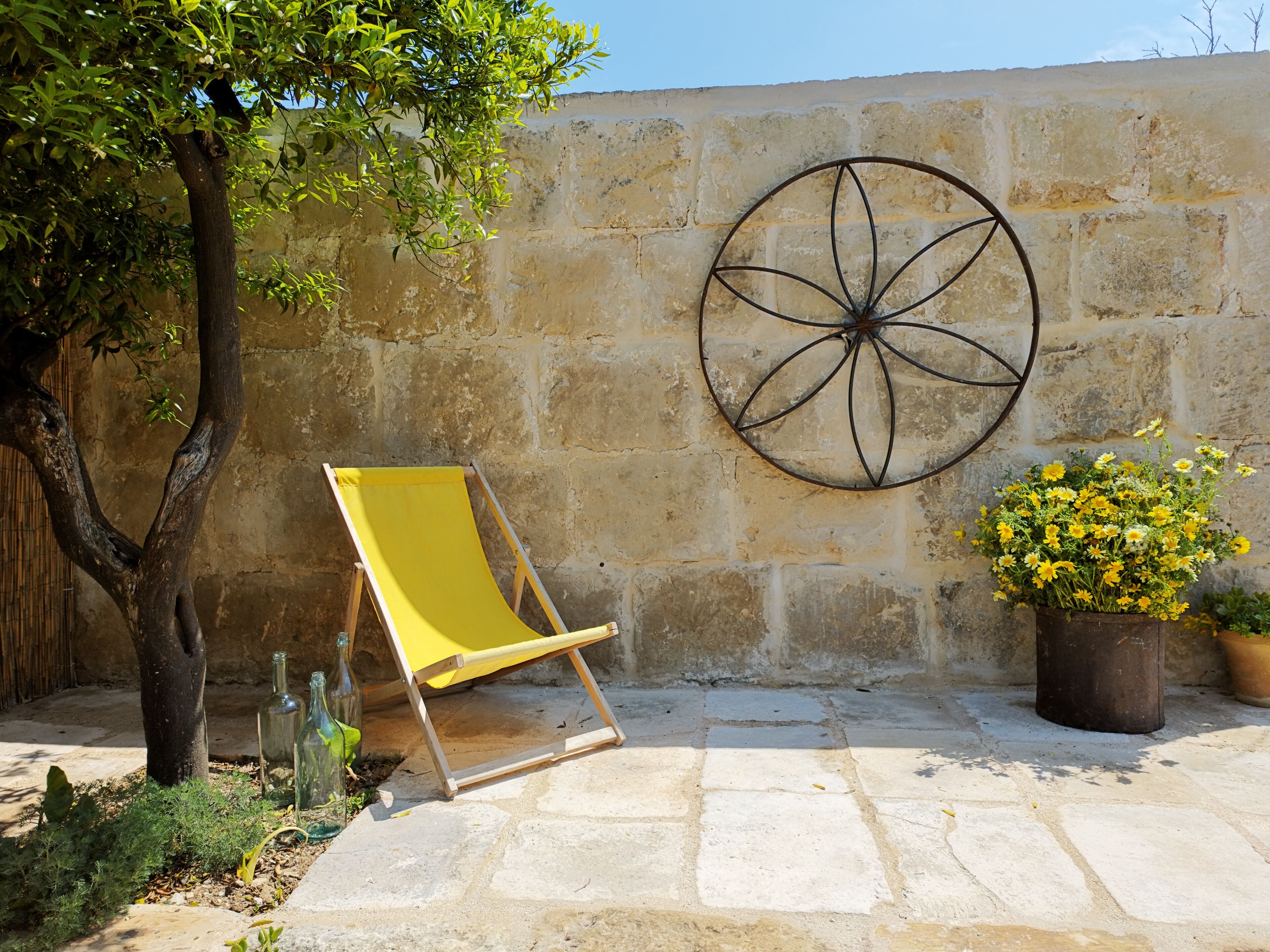 Una sedia da spiaggia gialla, un vaso con fiori gialli e una ruota decorativa in metallo su un muro di pietra.