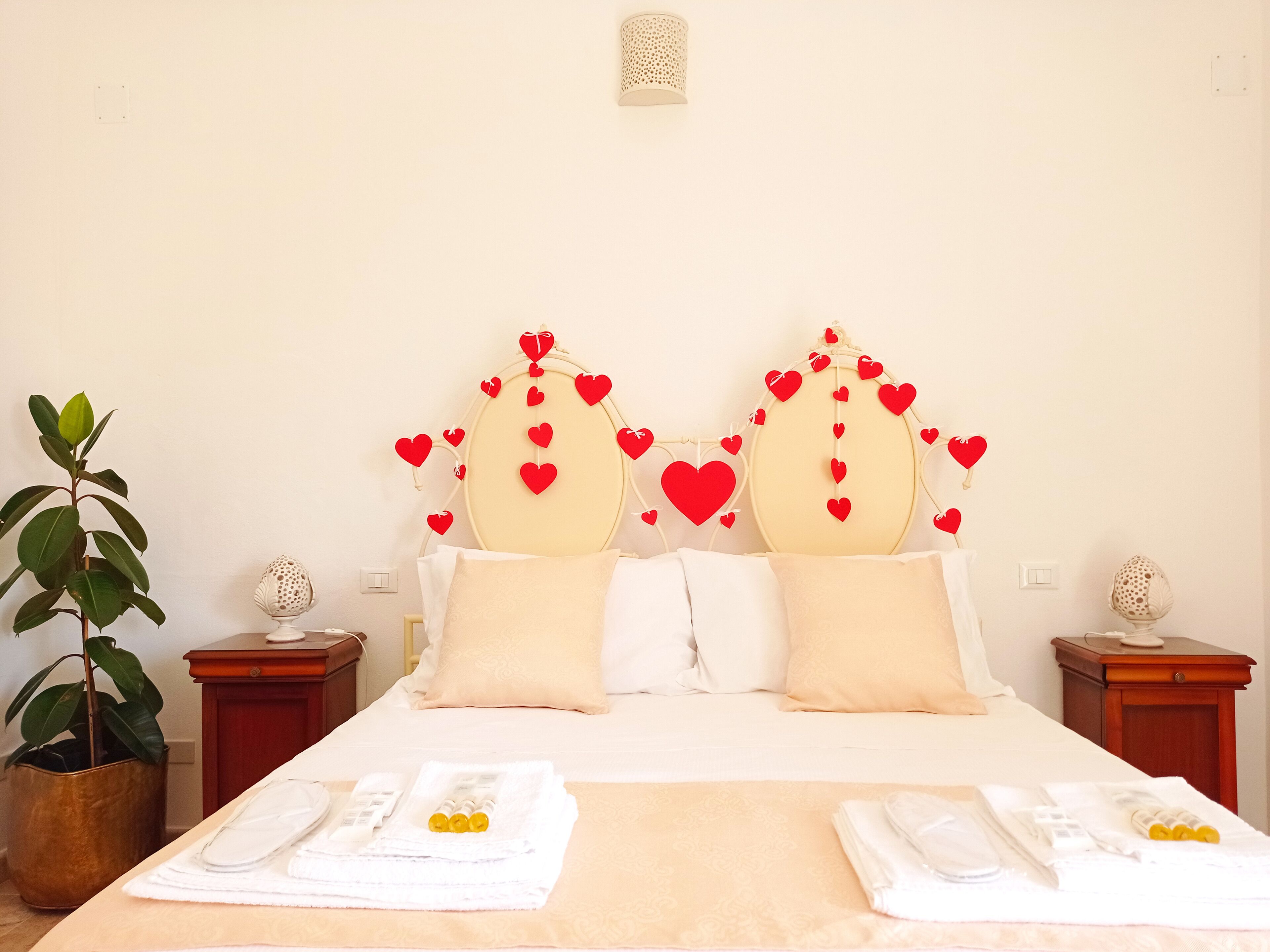 Une chambre d’hôtel avec un lit, deux tables de chevet, une plante en pot et des décorations en forme de cœur sur la tête de lit.
