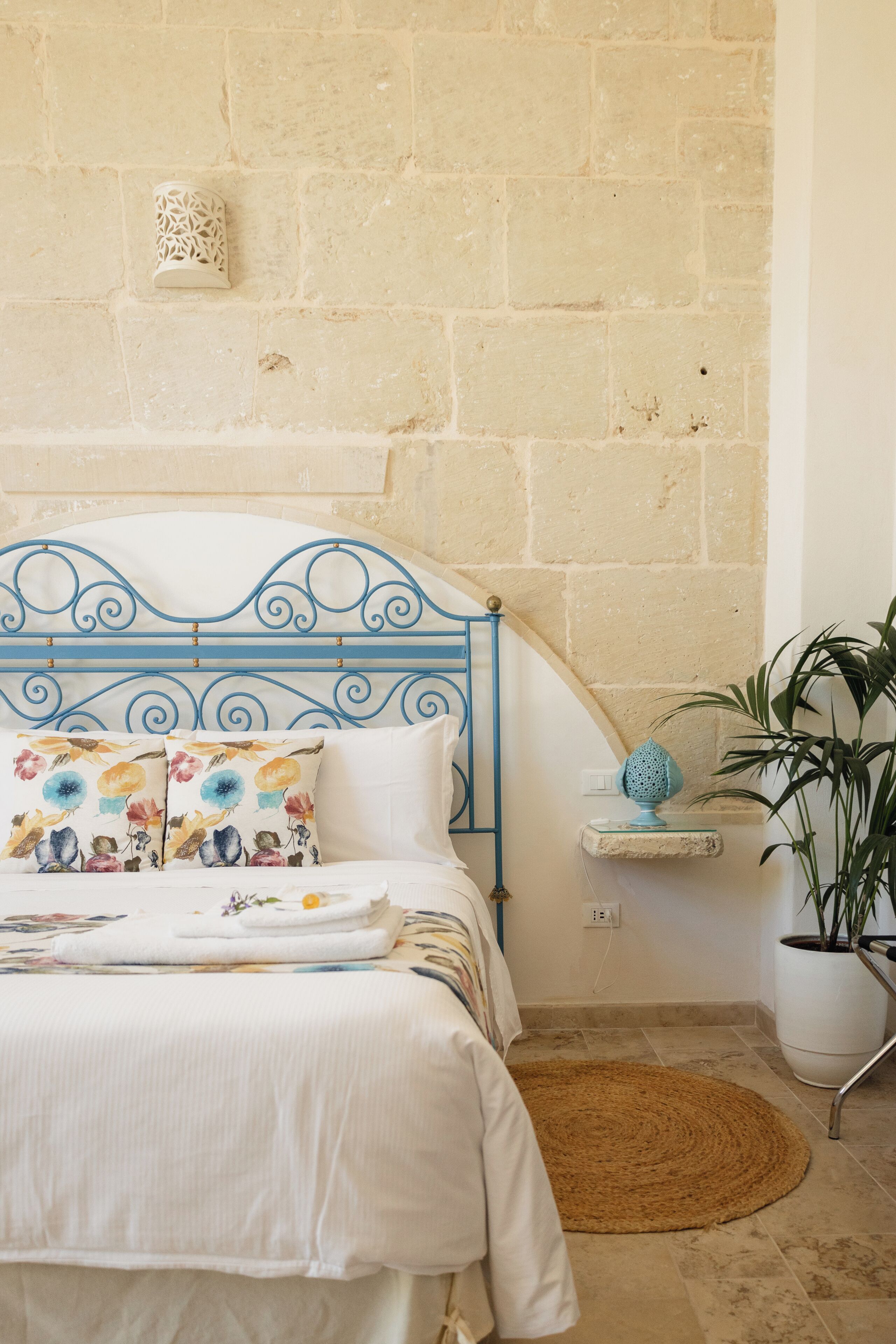 Uma cama com cabeceira de metal azul, um vaso com planta e luminária fixada na parede.
