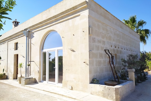 A stone building with a large arched entrance, a small balcony, and a door with a glass panel. - Giardino Frannicola (Maglie)