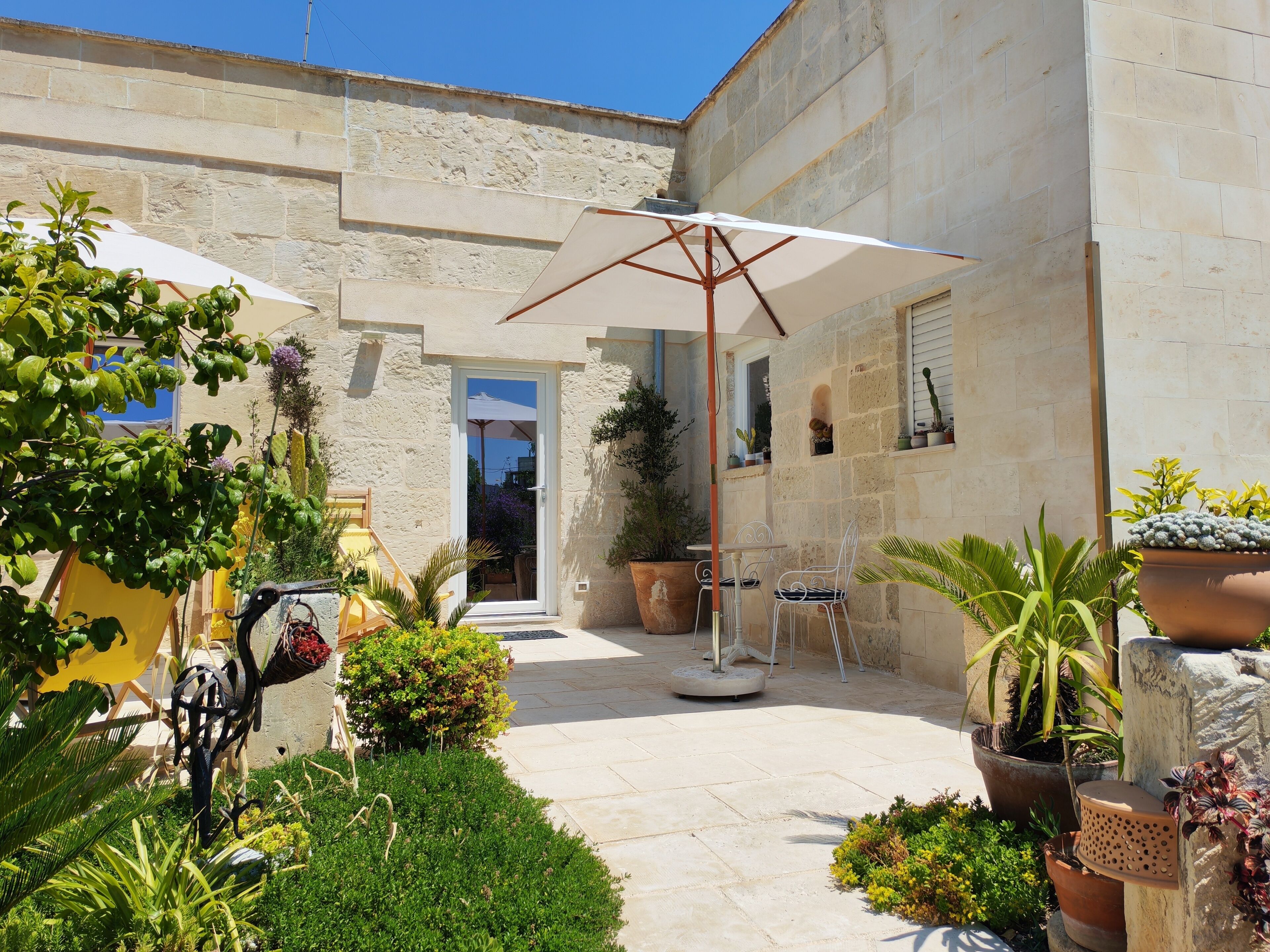 A courtyard with stone walls, potted plants, a white umbrella, and outdoor furniture.