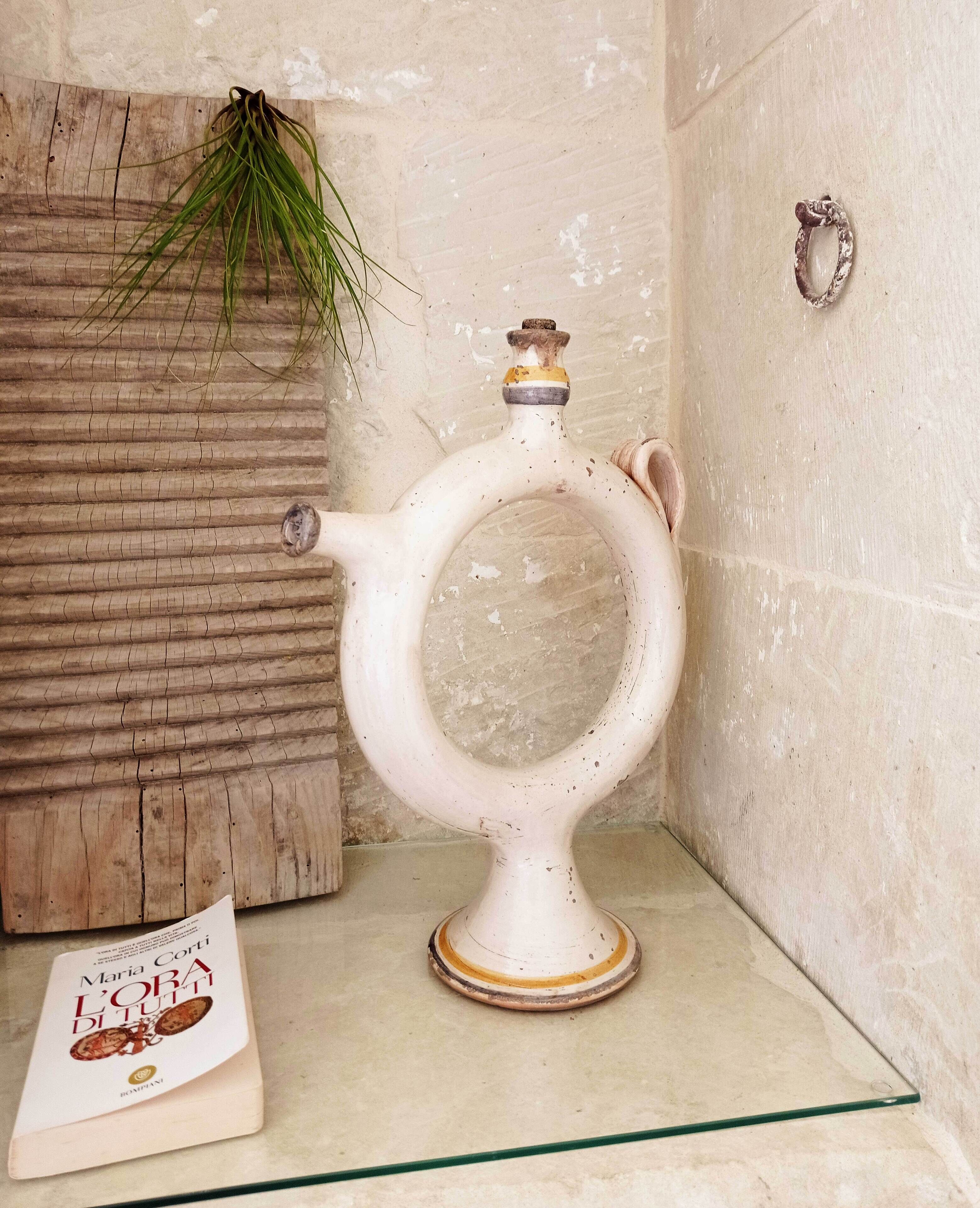 A white ceramic teapot with a handle and spout, placed on a glass shelf against a textured wall.