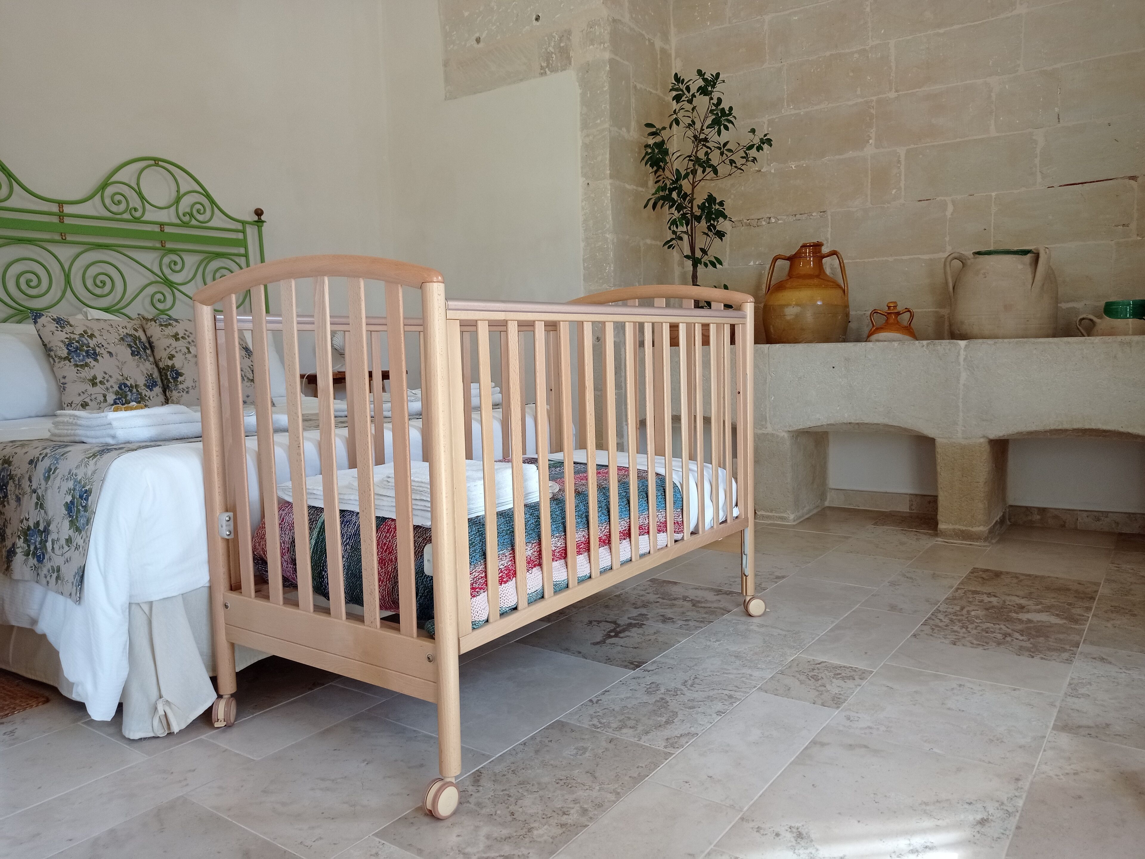 A wooden crib with a colorful quilt, a green metal headboard, a tiled floor, and a stone wall.