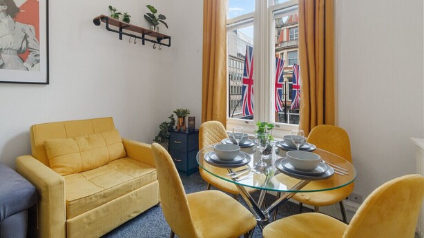 A room with a yellow sofa, a glass table set for dining, and a window with British flags. - Chic 1-Bedroom Retreat on Oxford Street - GC3 (Greater London)