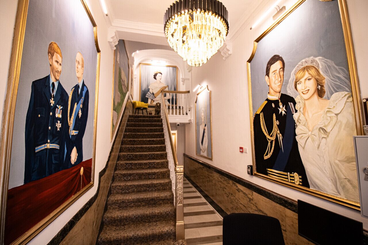 A grand hallway with a staircase, a chandelier, and two large framed portraits on the walls.
