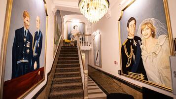 A grand hallway with a staircase, a chandelier, and two large framed portraits on the walls.