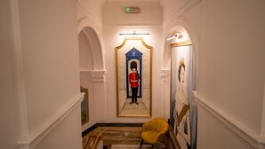 A hallway with a mirror reflecting a guard in uniform, a yellow chair, and a framed portrait. - Luxury at Your Doorstep: 2-Bedroom Gem on Oxford Street -10 (Greater London)