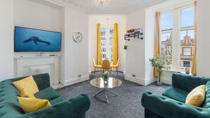 A living room with green sofas, a TV, a fireplace, a dining area with yellow chairs, and a view of buildings through large windows. - Luxury at Your Doorstep: 2-Bedroom Gem on Oxford Street -10 (Greater London)