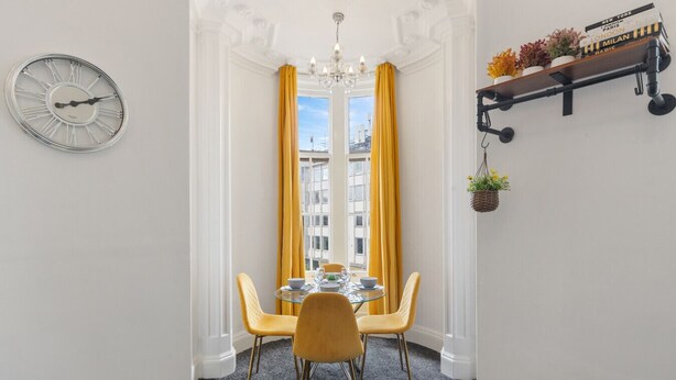 A dining area with a round table, yellow chairs, and a clock on the wall. - Luxury at Your Doorstep: 2-Bedroom Gem on Oxford Street -10 (Greater London)