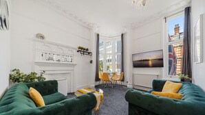 A room with two green sofas, a fireplace, a dining area with yellow chairs, and a TV on the wall.