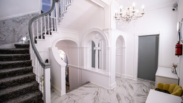 A grand staircase with marble steps and white walls, leading to an upper floor with a chandelier and a fire extinguisher on the wall.