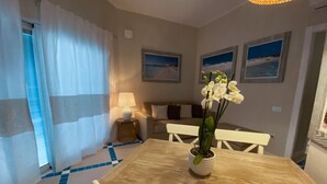 Un área de comedor con una mesa de madera, sillas blancas, una orquídea en maceta, una lámpara y un sofá con cojines.