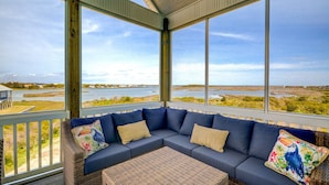 A covered porch with a blue sofa and wicker ottoman, overlooking a water body and grassy landscape. - Spacious 8-bedroom oceanview house in North Topsail Beach, perfect for gatherings! (North Topsail Beach)