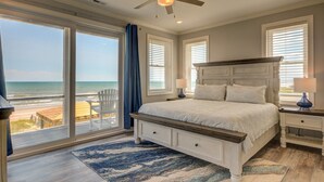 Een slaapkamer met een groot raam dat uitkijkt op het strand, een plafondventilator en een bed met een houten hoofdeinde.