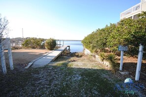 A wooden dock leading to a calm body of water, surrounded by greenery and a clear sky.