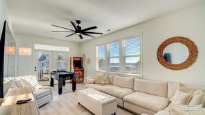 A spacious living room with a foosball table, a sofa, and a coffee table. There is a ceiling fan, large windows with blinds, and a round mirror on the wall. - Ocean Sounds: Spacious 7BR oceanview beach house in North Topsail with ocean views! (North Topsail Beach)