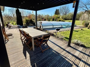 A covered outdoor patio with a table and chairs, a greenhouse, and a grassy area.