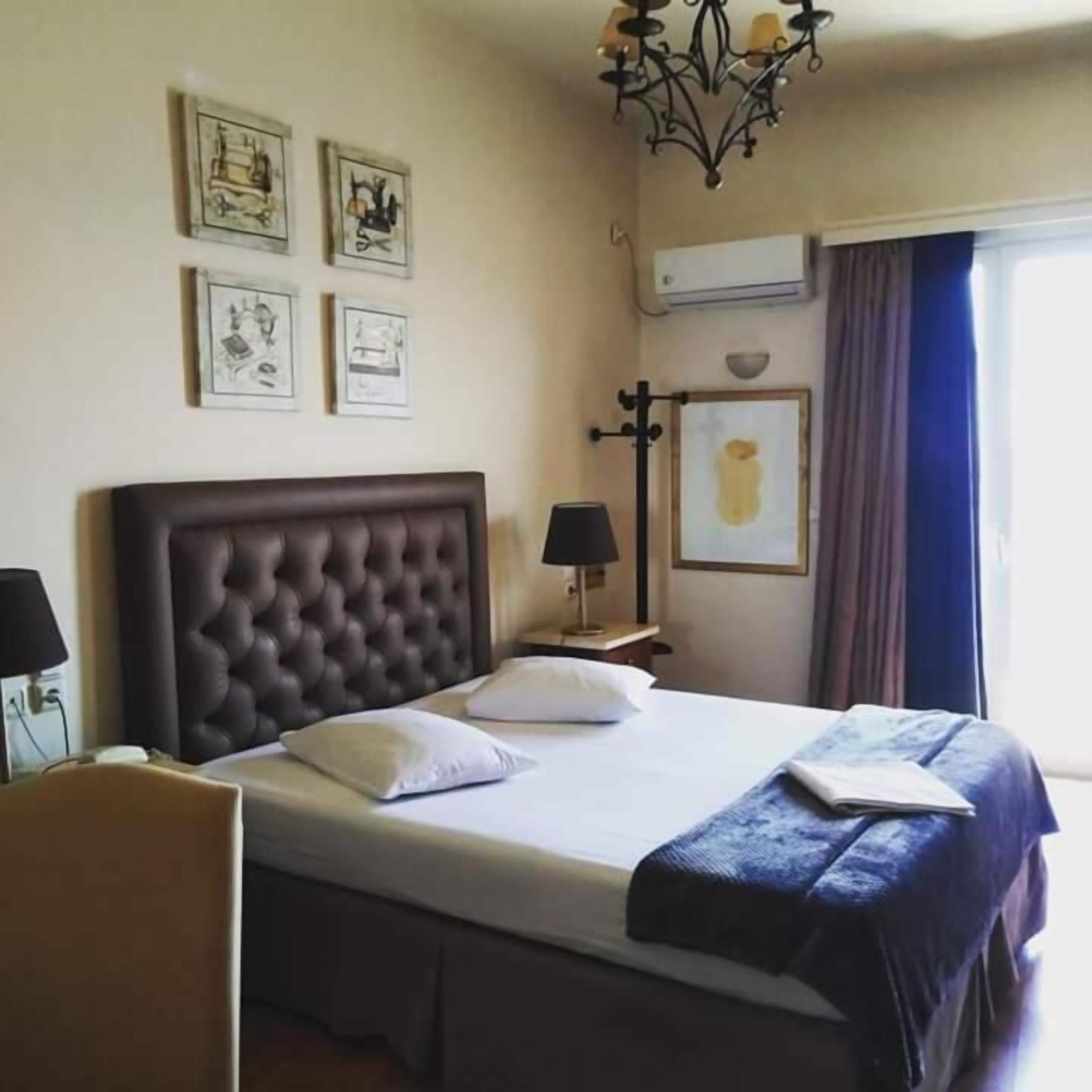 A hotel room with a large bed, a headboard, a bedside table with a lamp, a wall-mounted air conditioner, and framed pictures on the wall.