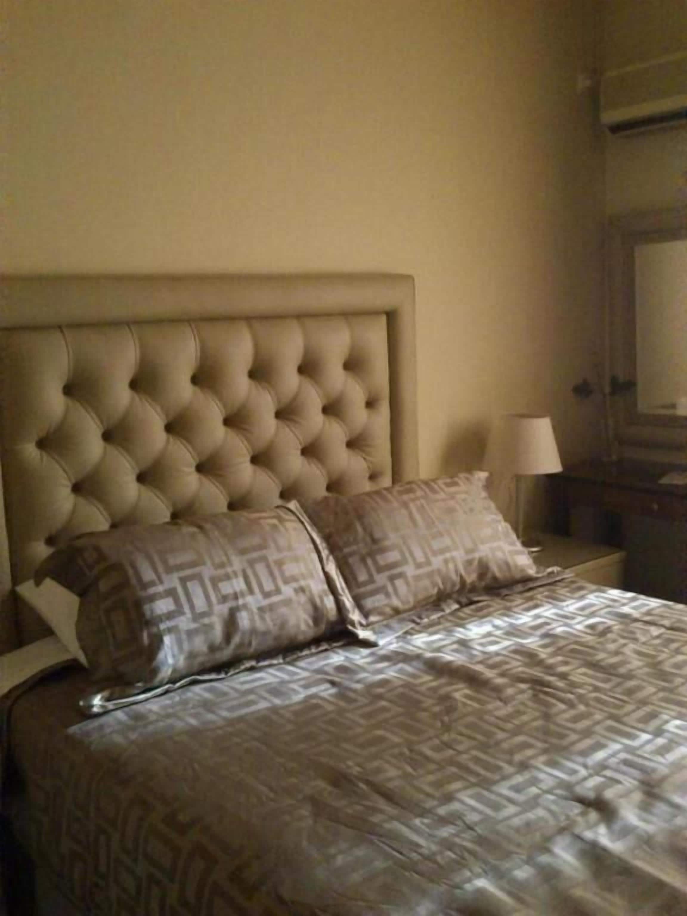 Uma cama com cabeceira estofada, duas almofadas estampadas e uma mesa de cabeceira com abajur.