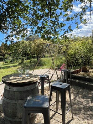 An outdoor seating area with a wooden table and stools, surrounded by greenery and trees.