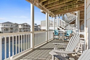 Une terrasse en bois, dotée de chaises et d’un escalier menant aux niveaux supérieurs, offre une vue sur une étendue d’eau et sur d’autres bâtiments.