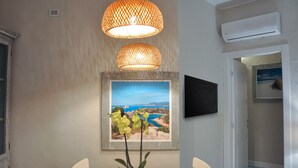 Un comedor con una mesa, sillas, una planta en maceta, un cuadro enmarcado de un paisaje costero y dos lámparas colgantes.