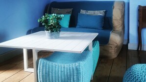 Een kamer met een blauwe bank, een witte tafel, een plant in pot en een blauwe geweven hocker.