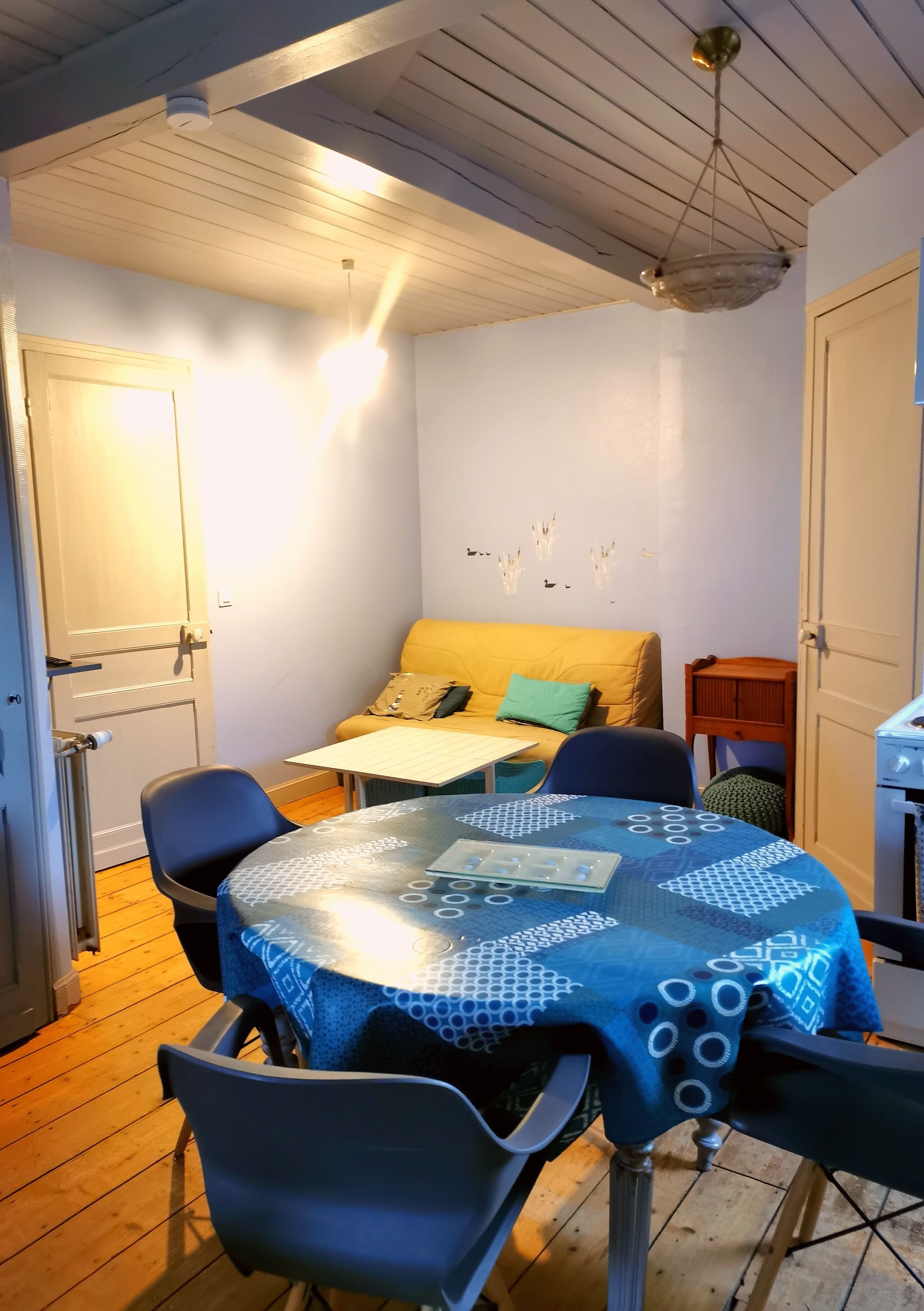 Ein kleiner, gemütlicher Raum mit Holzbalkendecke, einem Tisch mit blauer Tischdecke, einem gelben Sofa und einem kleinen Holzschrank.