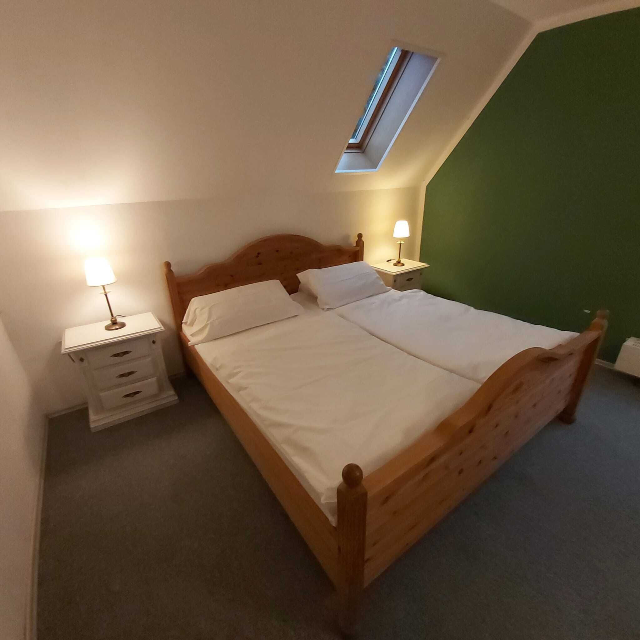 A bedroom with a wooden bed, two bedside tables with lamps, a small drawer unit, and a skylight.