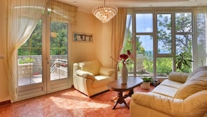 A spacious living room with a beige sofa, a small table with a vase, and a chandelier. There is a sliding glass door leading to an outdoor area with a balcony.