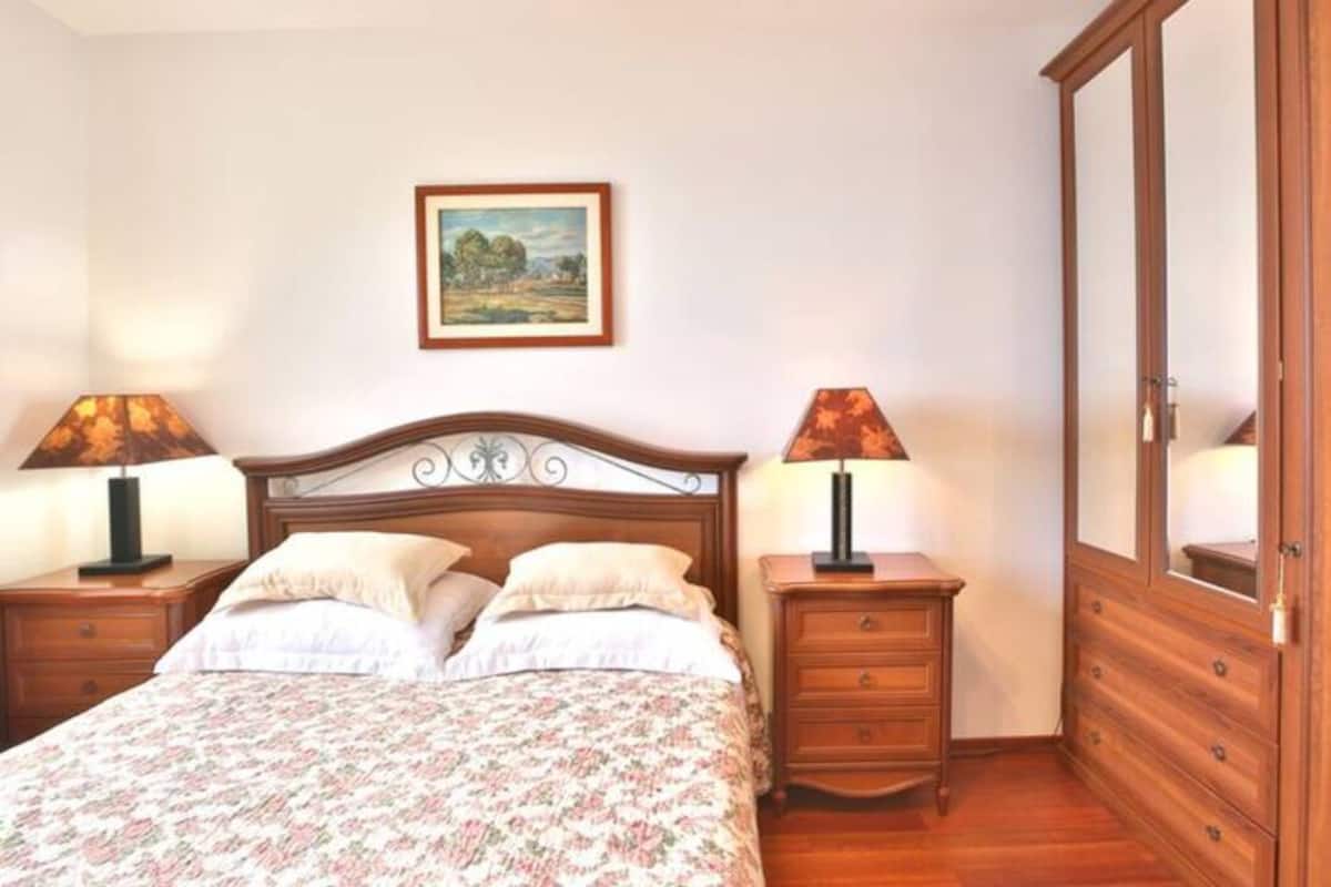 Une chambre à coucher avec un lit double, deux tables de chevet avec des lampes, une armoire en bois et un tableau représentant un paysage, accroché au mur.
