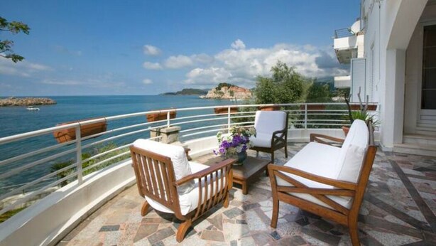 A balcony with wooden furniture, a flower vase, and a view of the sea and distant land.