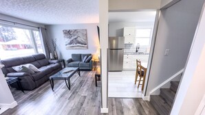 A modern living room with a grey sofa, a glass coffee table, and a painting on the wall. A kitchen with white cabinets and a stainless steel refrigerator is visible through an open doorway.