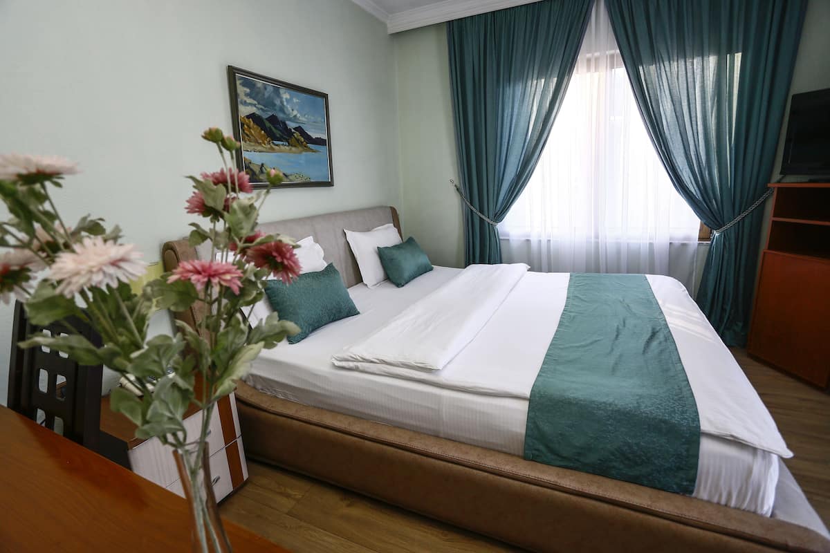 A neatly made bed with a white and teal bedspread, a brown headboard, and a teal pillow. A painting of a landscape is on the wall, and there is a vase with flowers on the table.