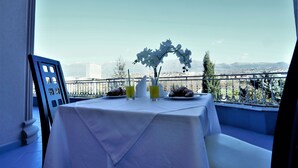 Una mesa con mantel blanco, dos vasos de zumo de naranja, un jarrón blanco con flores y dos cruasanes en platos, con vistas a un paisaje urbano con montañas al fondo.
