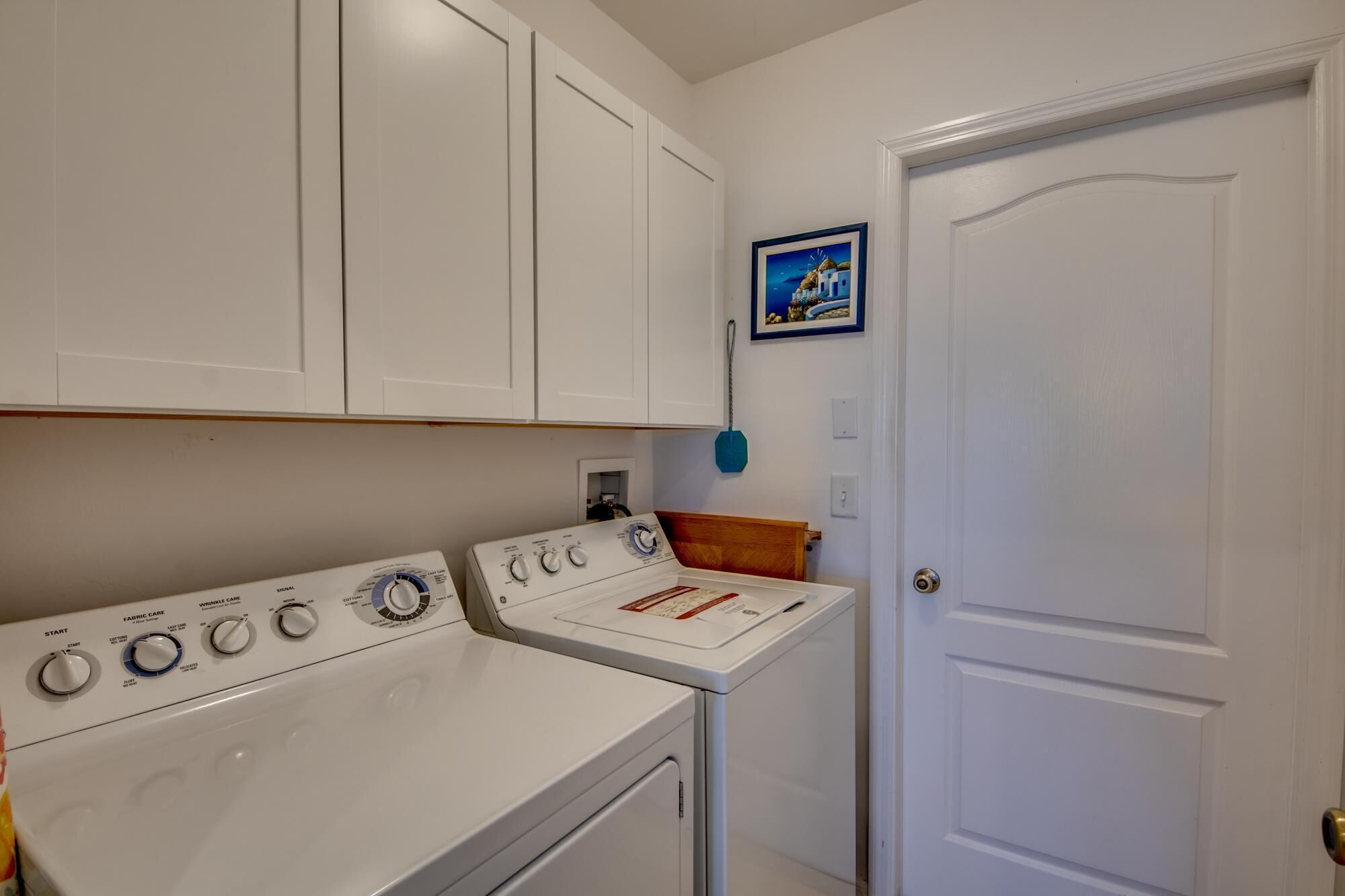 A laundry room with a white washer and dryer, a wooden cabinet, a door, and a small framed picture on the wall.