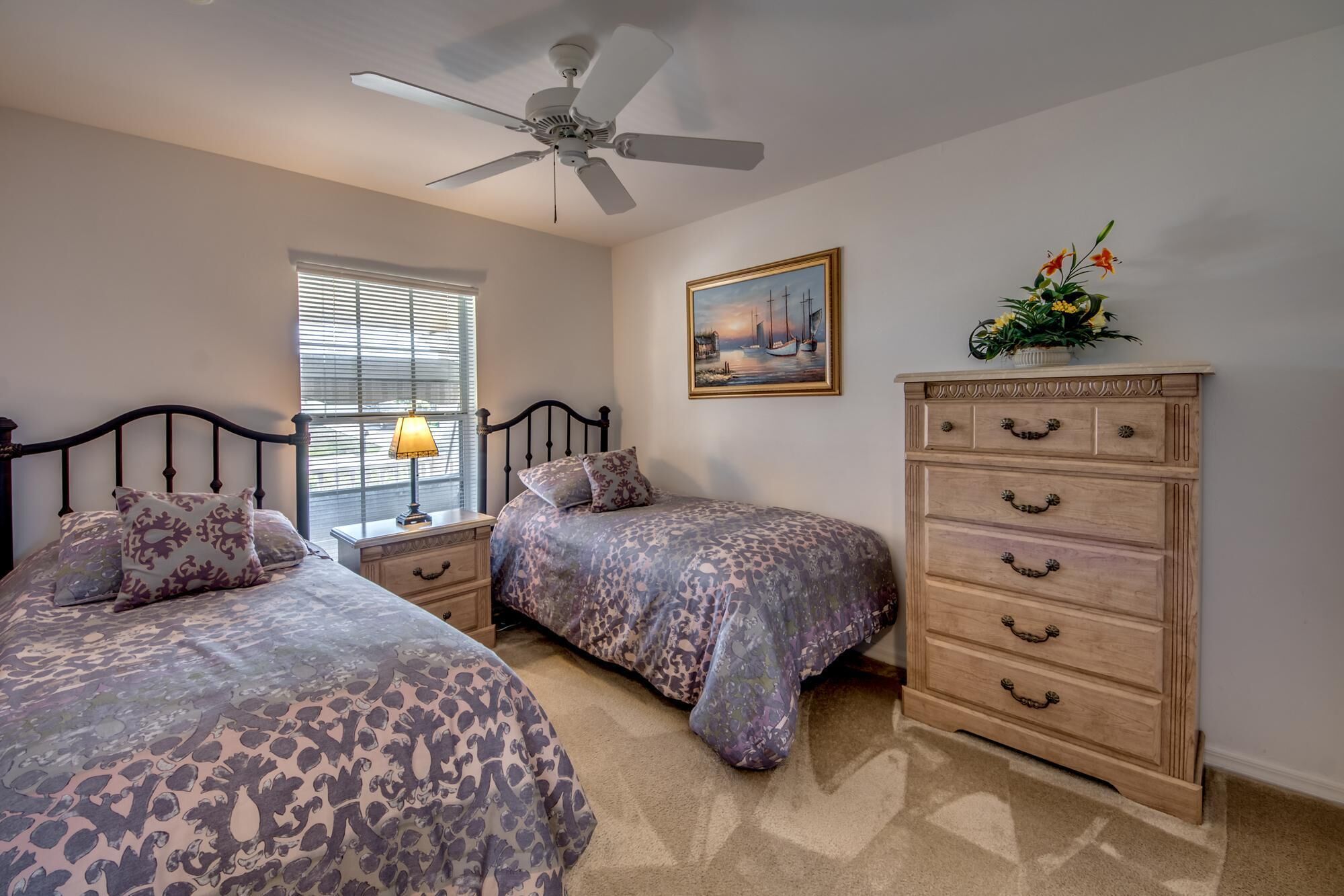 A bedroom with two beds, a ceiling fan, a window with blinds, a nightstand with a lamp, a dresser with drawers, and a painting on the wall.