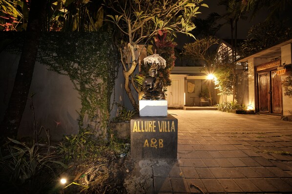 A nighttime view of a villa entrance with a stone sign reading "Allure Villa A&B" and a statue on top.