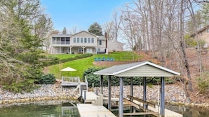 Uma casa com um píer que se estende sobre um lago, rodeada por árvores e um gramado bem cuidado.