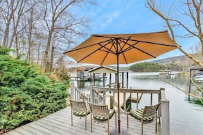 A wooden deck with outdoor seating and a large umbrella, overlooking a lake with boats and a dock.