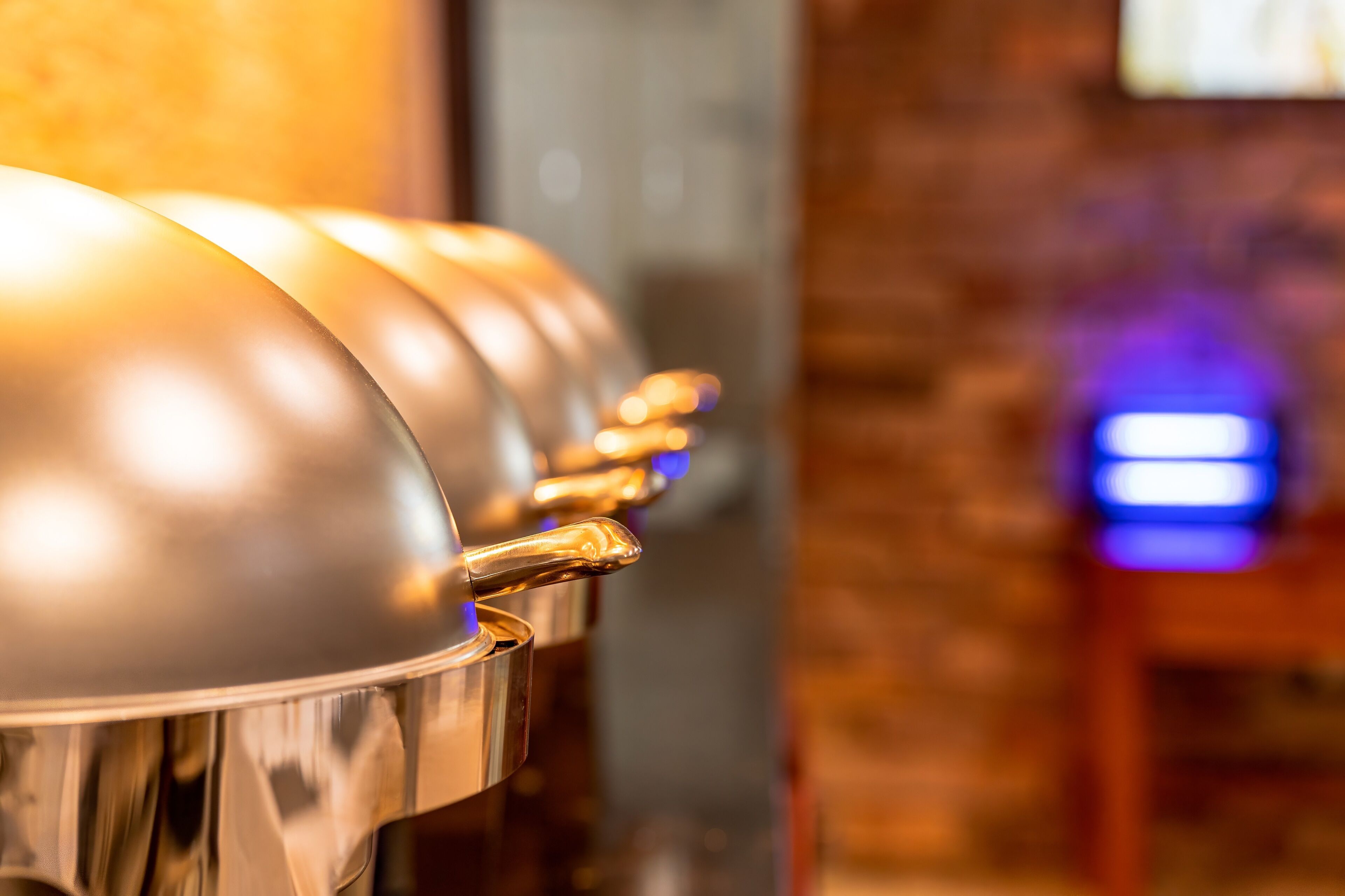 A row of metallic, reflective food service equipment, likely for a buffet or catering service, in a setting with a brick wall and a blue-lit area in the background.