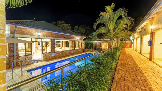 A poolside area with a brick building, a swimming pool, and a pathway leading to a covered seating area. - The Shrub lodge (Victoria Falls)